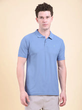 Man wearing a blue polo t-shirt  (DCYPHR Men's Element Polo - Denim Blue) against a beige background front facing towards camera.